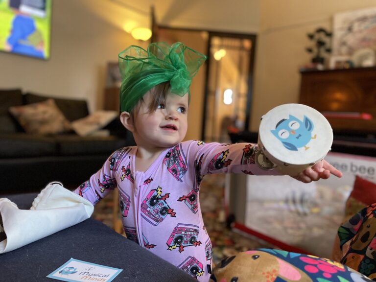 Toddler wearing a green headwrap and pink pajamas holding a tambourine with a blue owl illustration.