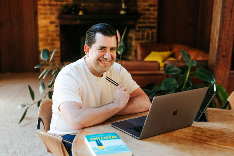 Smiling man in white shirt seated at wooden table with laptop, pen, and book.