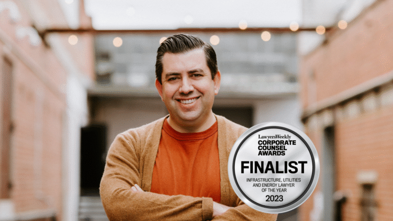 Smiling man in brown cardigan and orange shirt with 2023 Corporate Counsel Awards finalist badge.