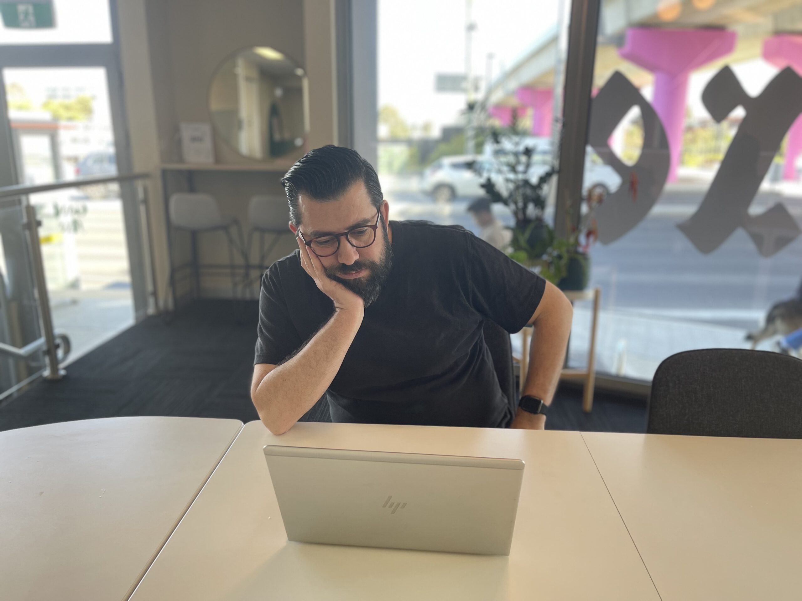Man with glasses and beard looking at laptop in modern office with large windows.