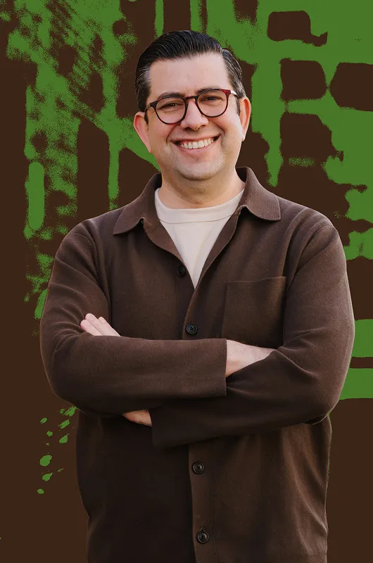 Portrait of a smiling man with glasses wearing a brown jacket and beige shirt.