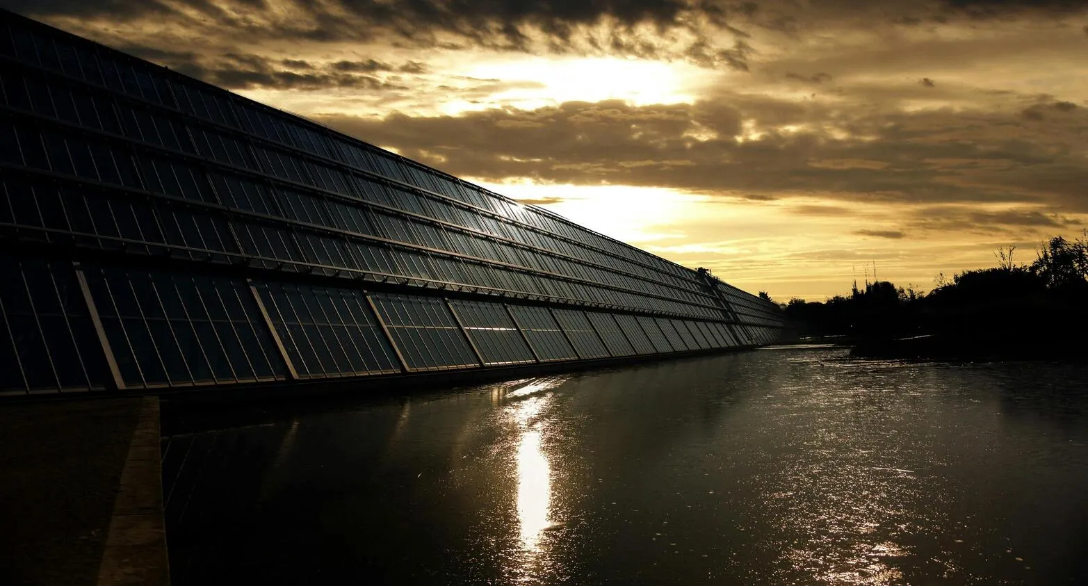 Solar panels reflecting sunlight at sunset beside a calm body of water.
