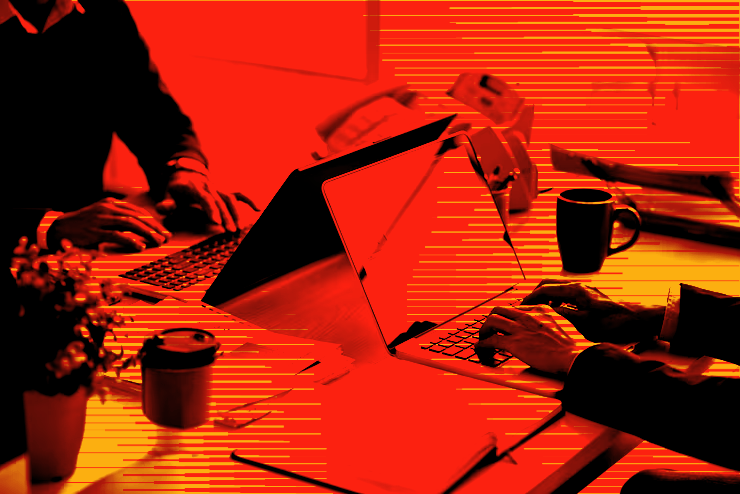 Two individuals working on laptops at a desk with coffee cups and office supplies.